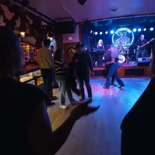 People dancing and socializing in a dimly lit venue with a live band performing on stage under blue and purple lighting